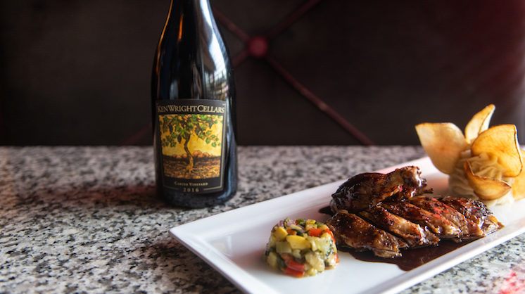 A dish of cooked meat and vegetables served in a cylinder shape sits next to a bottle of red wine.