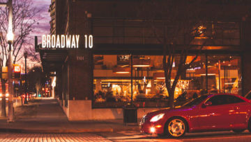 The brick exterior of Broadway 10 on Automobile Alley in Oklahoma City. The sky is a purple-yellow dusk and a red car is pulled up to a stoplight.
