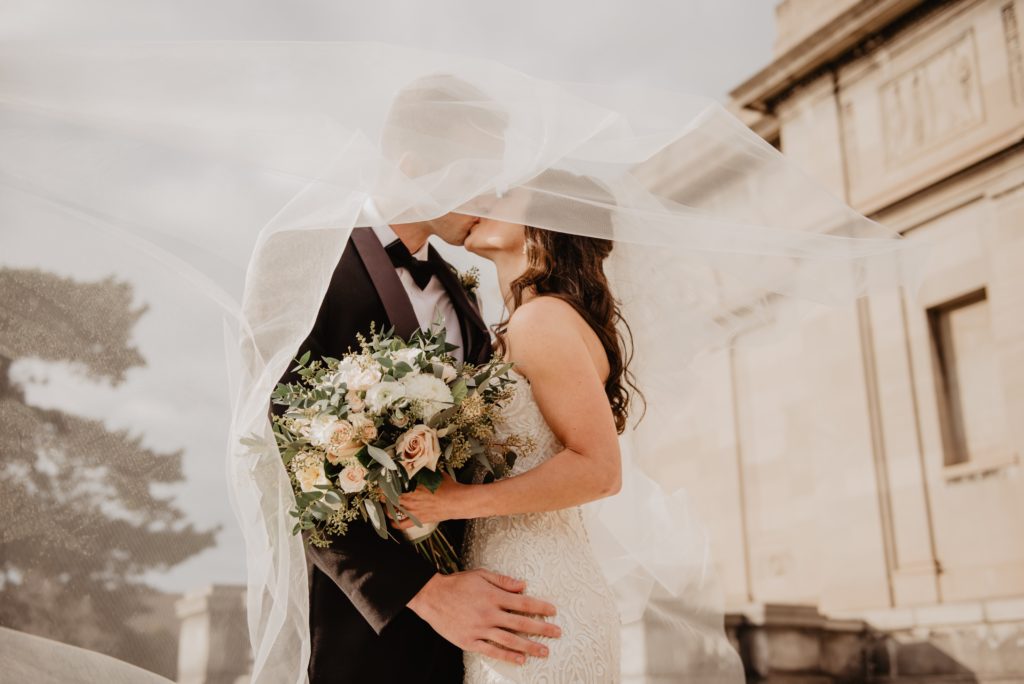 A bride and groom kiss underneath the bride's veil that's twirling in the wind.