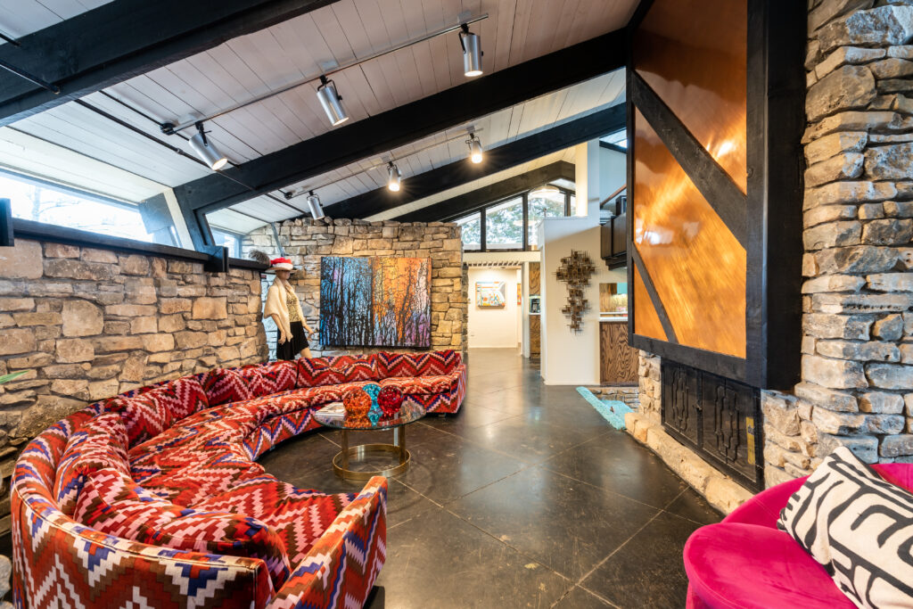 An open living room in a mid-century modern home in the Quail Creek Home Tour, with an angled roof, stone walls and an arched red patterned couch.