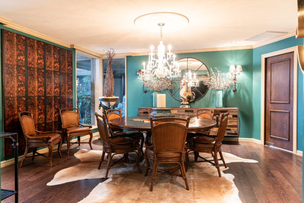 A dining room with green textured wallpaper, dark wood table and chairs and a skinned rug at the Quail Creek Home Tour. An elaborate chandelier hangs above and washes the room in yellow light. 