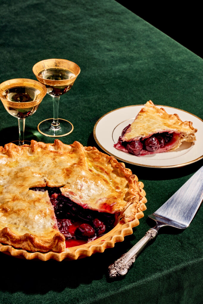 Cherry pie next to gold rimmed glasses on a green velvet tablecloth