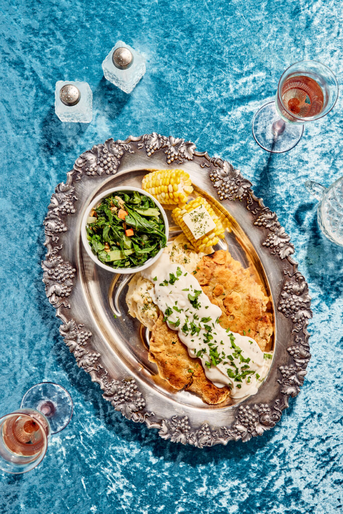 Birds eye view of a chicken fried steak with corn and veggies on silver serving dish sitting on blue velvet tablecloth