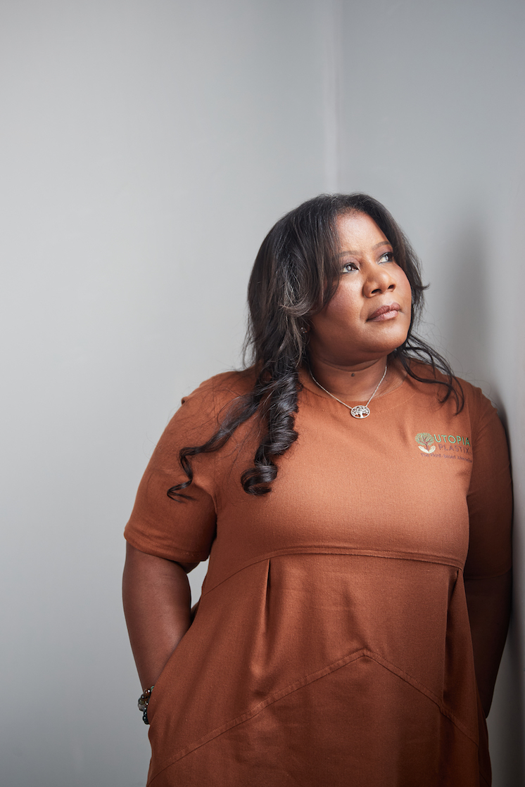 Sharina Perry of Utopia Plastix wears a brown dress with her company's logo and looks up while leaning against a wall.